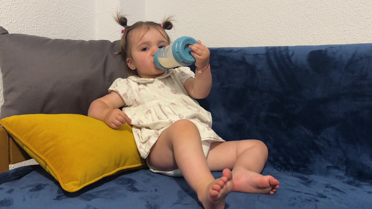 Bambino che beve dal biberon mentre si rilassa sul divano di casa, su un divano imbottito in feltro con un cuscino giallo