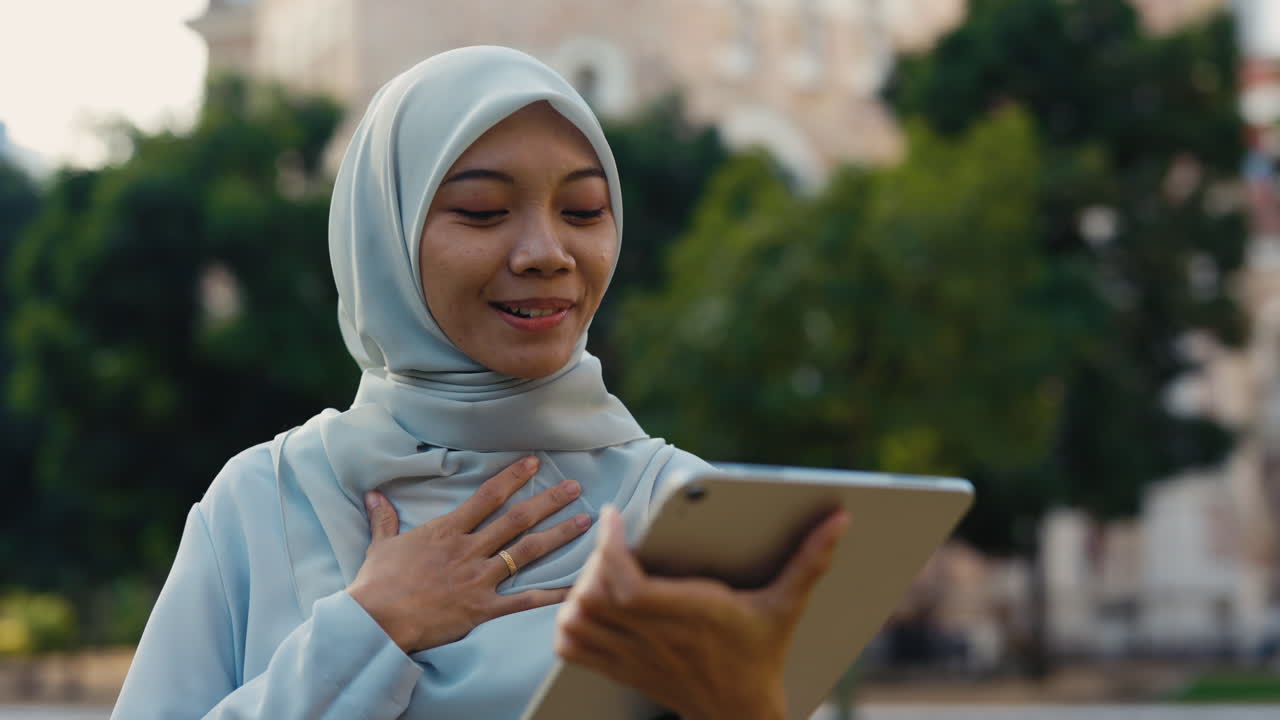 Young Muslim woman in hijab smiles during a video call on a tablet outdoors