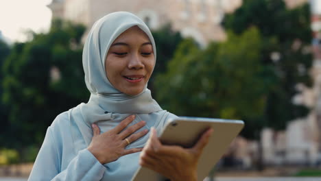 young muslim woman in hijab smiles during a video call on a tablet outdoors