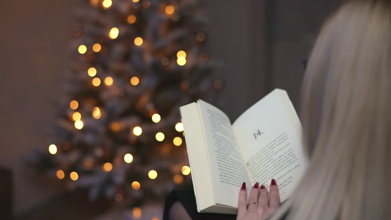 A woman with long hair is reading a book while sitting near a beautifully lit Christmas tree. The warm bokeh lights in the background create a peaceful and cozy holiday atmosphere