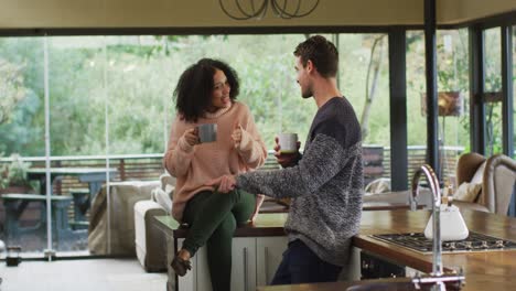 Happy-diverse-couple-talking-and-drinking-coffee-in-kitchen,-woman-sitting-on-counter