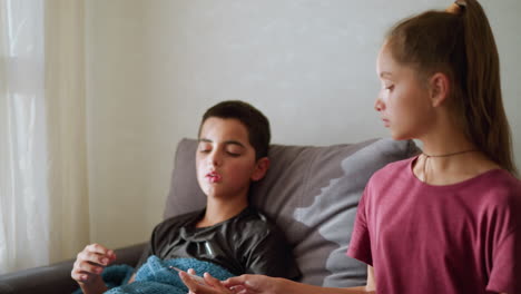 girl with rope around her neck hands thermometer to sick brother sitting on couch to check his temperature as he places it under his armpit, soft light filters through window