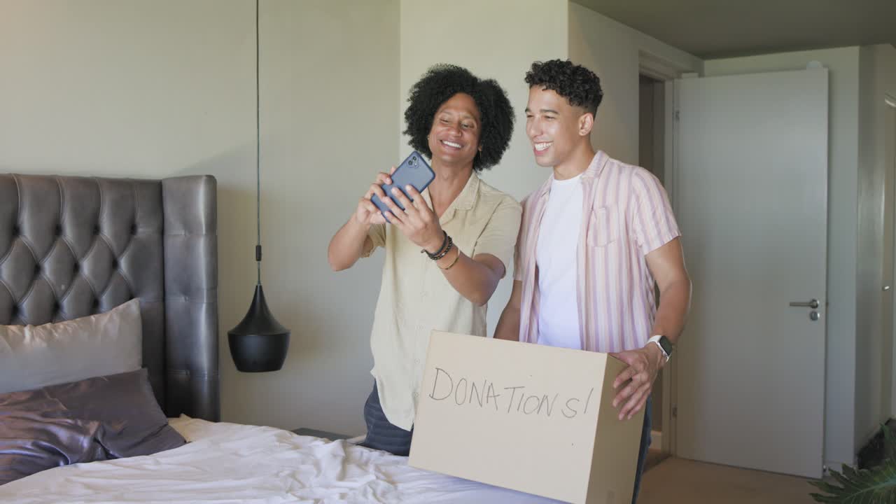 Amigos afro-americanos separando roupas no quarto, embalando caixa de DOAÇÕES!, verificando o celular, posando