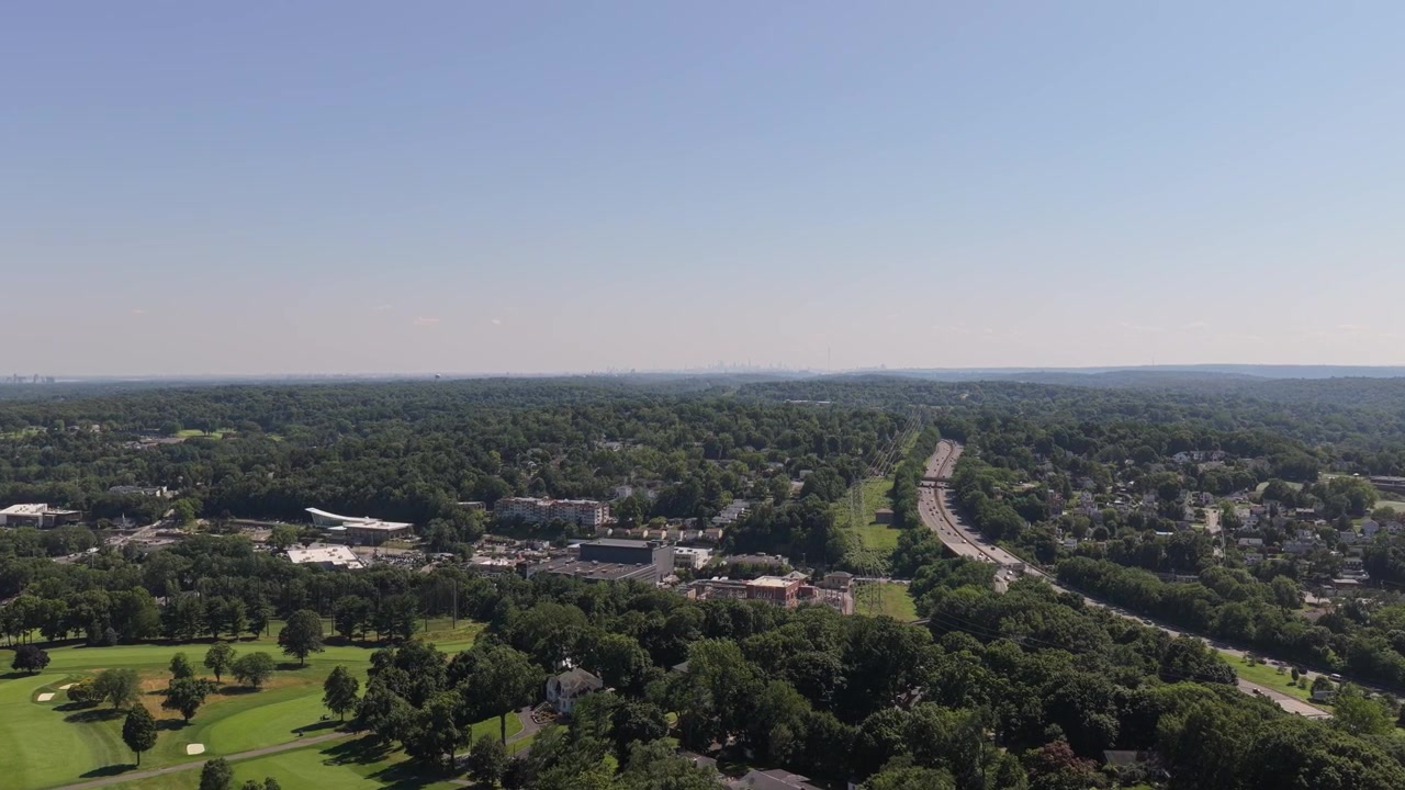 Ampia prospettiva aerea che sorvola un paesaggio verde lussureggiante con un campo da golf e una città suburbana, muovendosi verso un'autostrada trafficata a più corsie sotto un cielo azzurro e limpido in estate