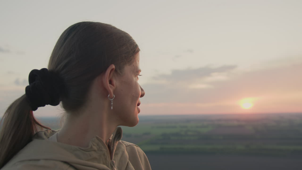Side view of woman in beige jacket gazing at glowing sunset while floating in hot air balloon, surrounded by expansive farmland below and gentle clouds above
