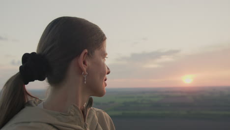 side view of woman in beige jacket gazing at glowing sunset while floating in hot air balloon, surrounded by expansive farmland below and gentle clouds above