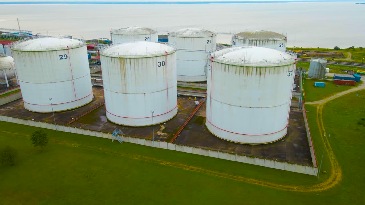 Aerial drone footage revealing large containers or tanks at a cloudy harbor made for storaging liquid gas or oil. The large tanks are numbered and baltic sea is visible in the horizon at Paldiski