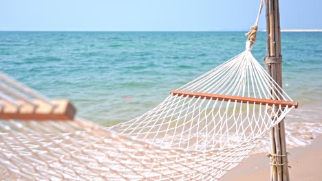 An-empty-rope-hammock-hangs-over-a-beach,-and-the-incoming-tide-while-it-rocks-in-the-breeze