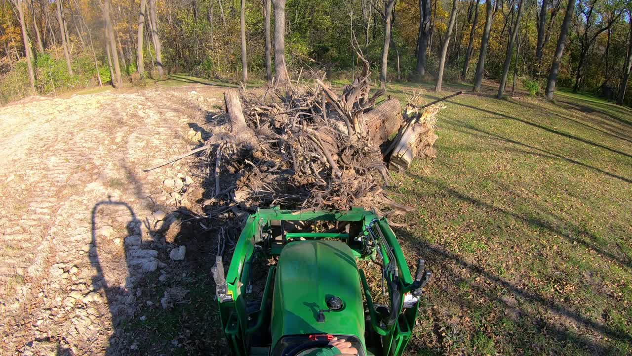 punto de vista de ángulo alto en un pequeño tractor verde usando horquillas elevadoras para recoger un tocón de árbol de un montón y mover escombros cerca de los bosques a principios de otoño