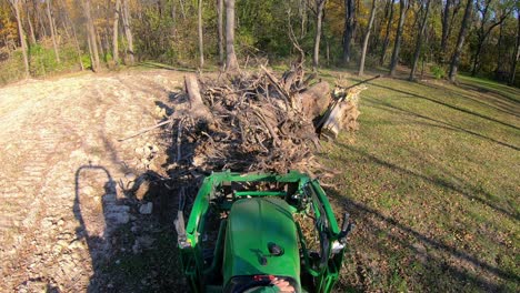 High-angle-point-of-view-on-small-green-tractor-using-lift-forks-to-pickup-a-tree-stump-from-a-pile-and-move-debris-near-woods-in-early-autumn