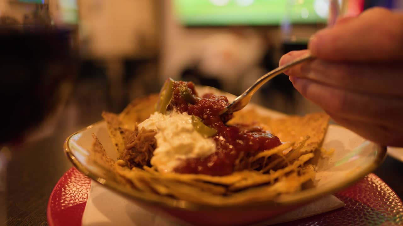 Dettaglio di nachos presi con una forchetta in un bar al chiuso con illuminazione calda e soffusa, messa a fuoco parziale