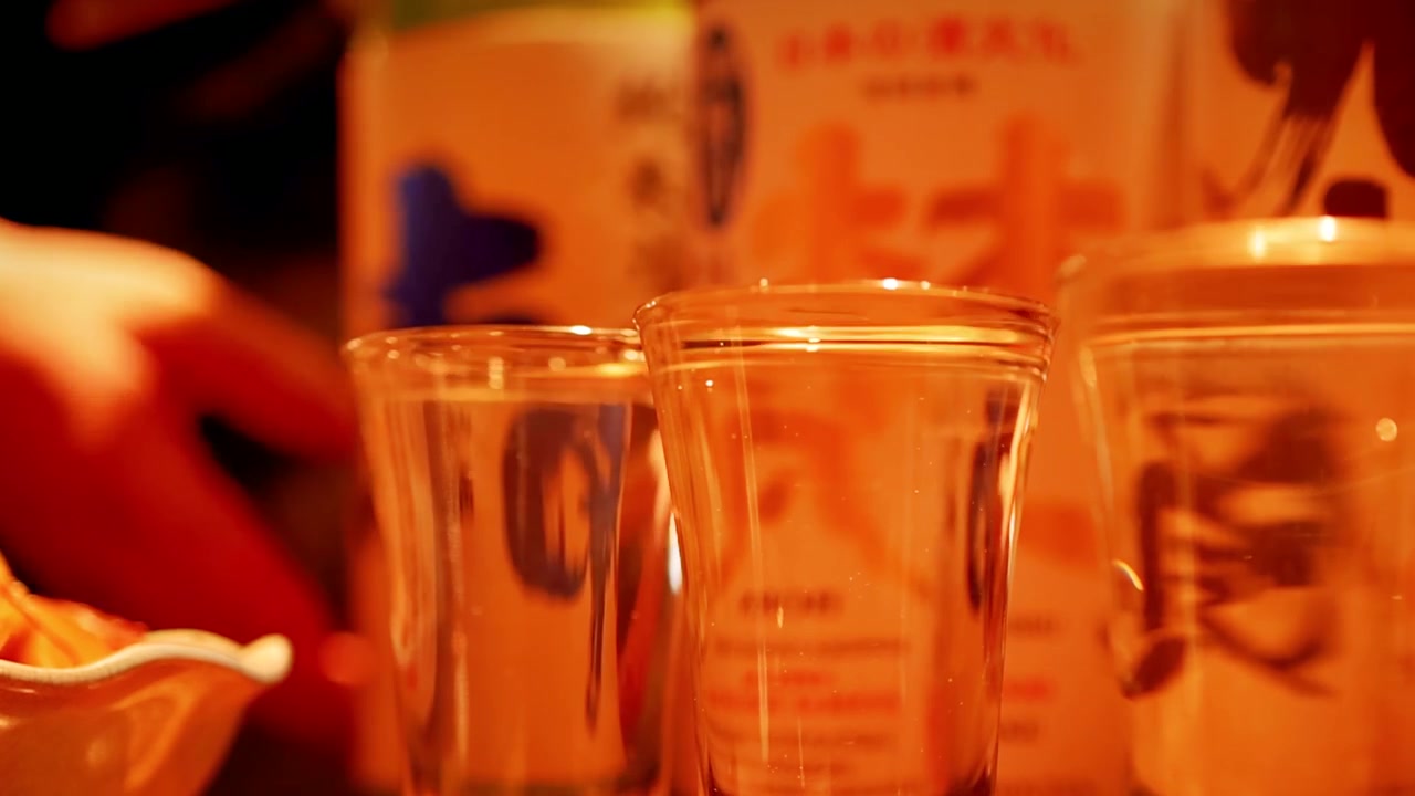 Sake bening dituangkan ke dalam gelas kecil di suasana restoran Jepang yang hangat dan remang-remang.