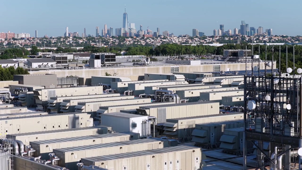 Rækker af industrielle køleenheder dækker et stort datacenter-tag i Secaucus, New Jersey, fjerne One World Trade Center rejser sig bag en tæt grøn trækant under blå himmel. Bredt dronebillede fra luften