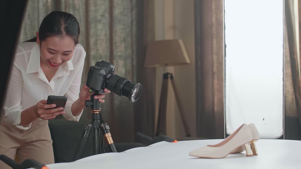 Asian Female Photographer Comparing Photos On A Smartphone To Camera While Taking Photos Of Women'S Shoes In Home Studio