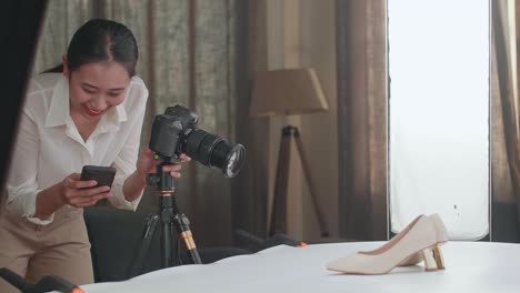 asian female photographer comparing photos on a smartphone to camera while taking photos of women's shoes in home studio