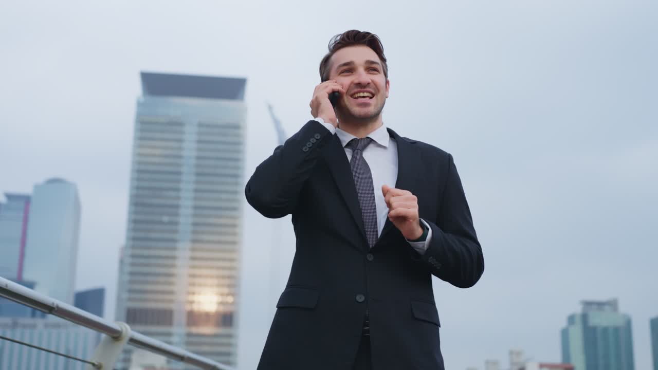 Empresario sonriente en traje se da la vuelta mientras mantiene una conversación telefónica con los imponentes edificios de la ciudad de Yeouido en Corea del Sur al atardecer: toma de paralaje en ángulo bajo