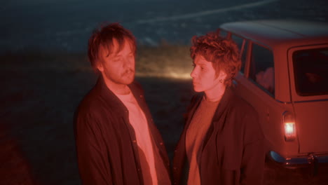 pensive couple standing under red light near vintage car at night