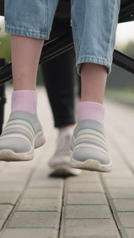 legs of schoolgirl with spinal injury sitting in wheelchair. mother pushes medical equipment with daughter walking on pavement in park closeup