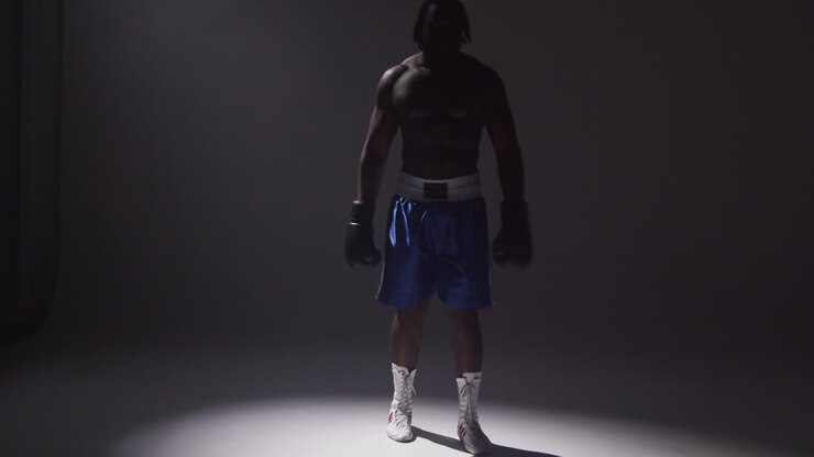 Toma de estudio silueteada con iluminación de clave baja de boxeador con guantes de pie en un haz de luz sobre fondo gris 2