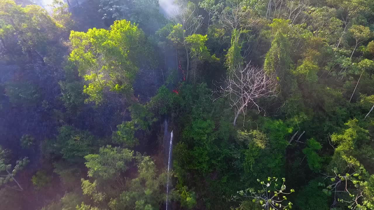 vista aérea de un bombero rociando agua de su manguera de escalera sobre el humo que se eleva, de un incendio forestal brillante - seguimiento, disparo de dron
