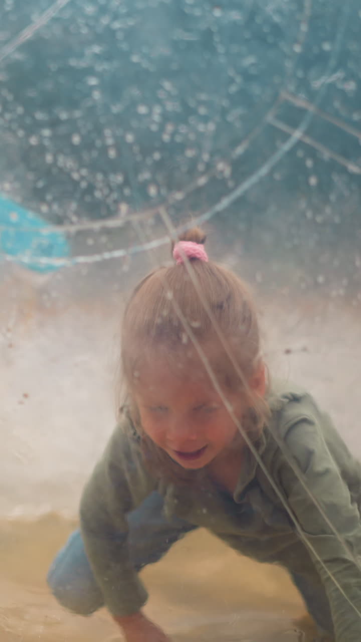 Happy little girl with braid crawls inside large transparent water ball floating in small pool on gloomy spring day closeup slow motion