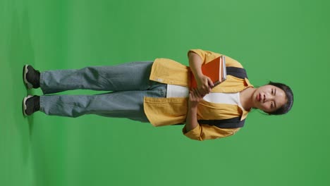 full body of unsatisfied asian woman student with a backpack and some books looking at the camera and shaking her head while standing in the green screen background studio