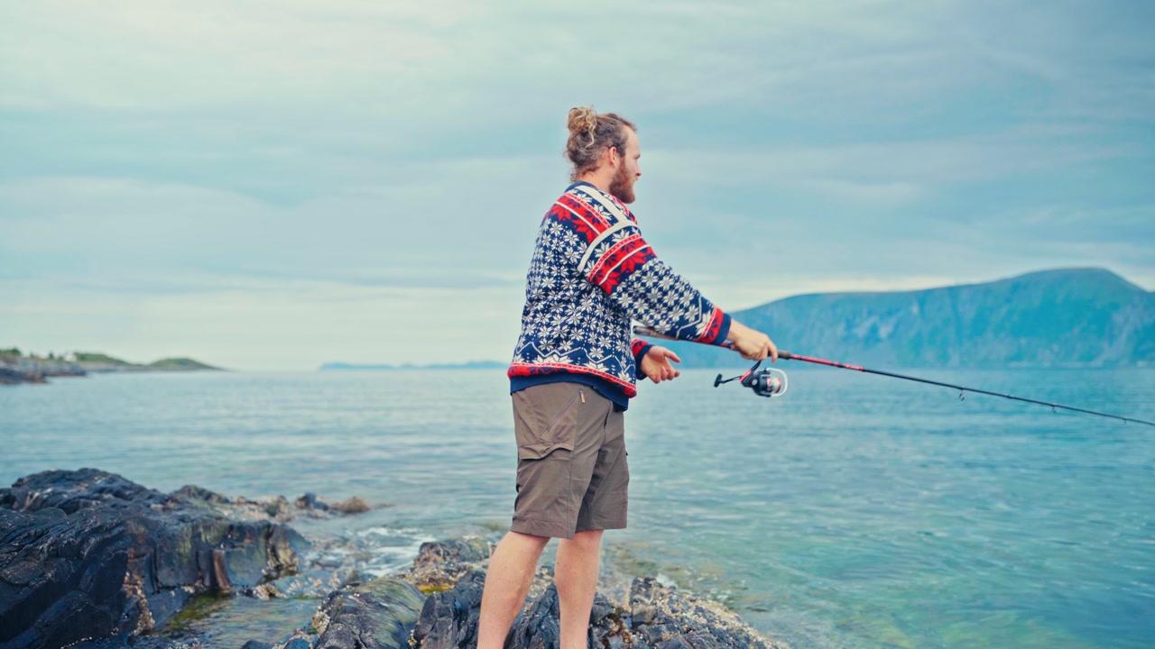 A Man Enjoys Fishing by the Shore, With Serene Waters and Distant Mountains in Sandland, Loppa Municipality, Norway - Close Up