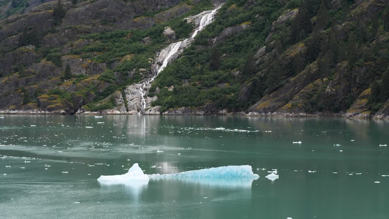Picturesque waterfall along the granite steep wall of the mountain and icebergs floating at Endicott Arm fjord, Alaska.