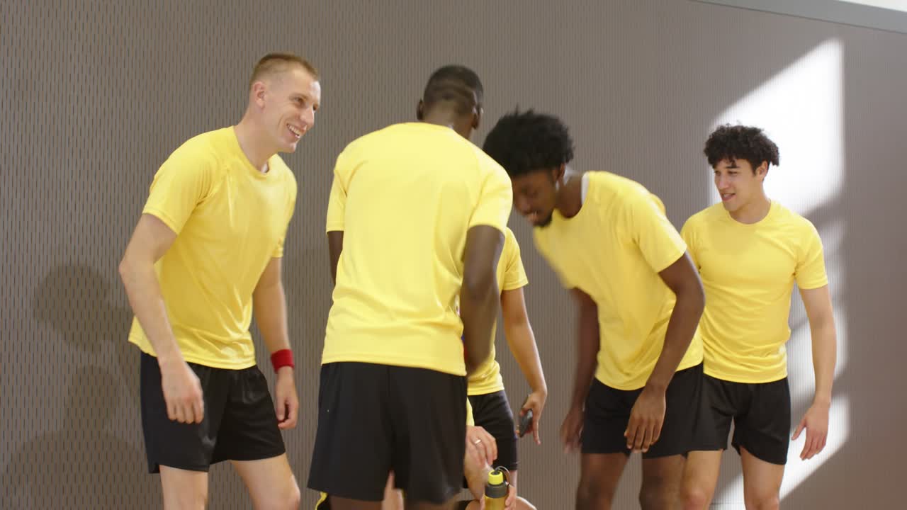 Ett volleybollag bestående av män med olika bakgrunder, klädda i gult, närmar sig bänken och hälsar, passar bollen i gymmet under hejarop.