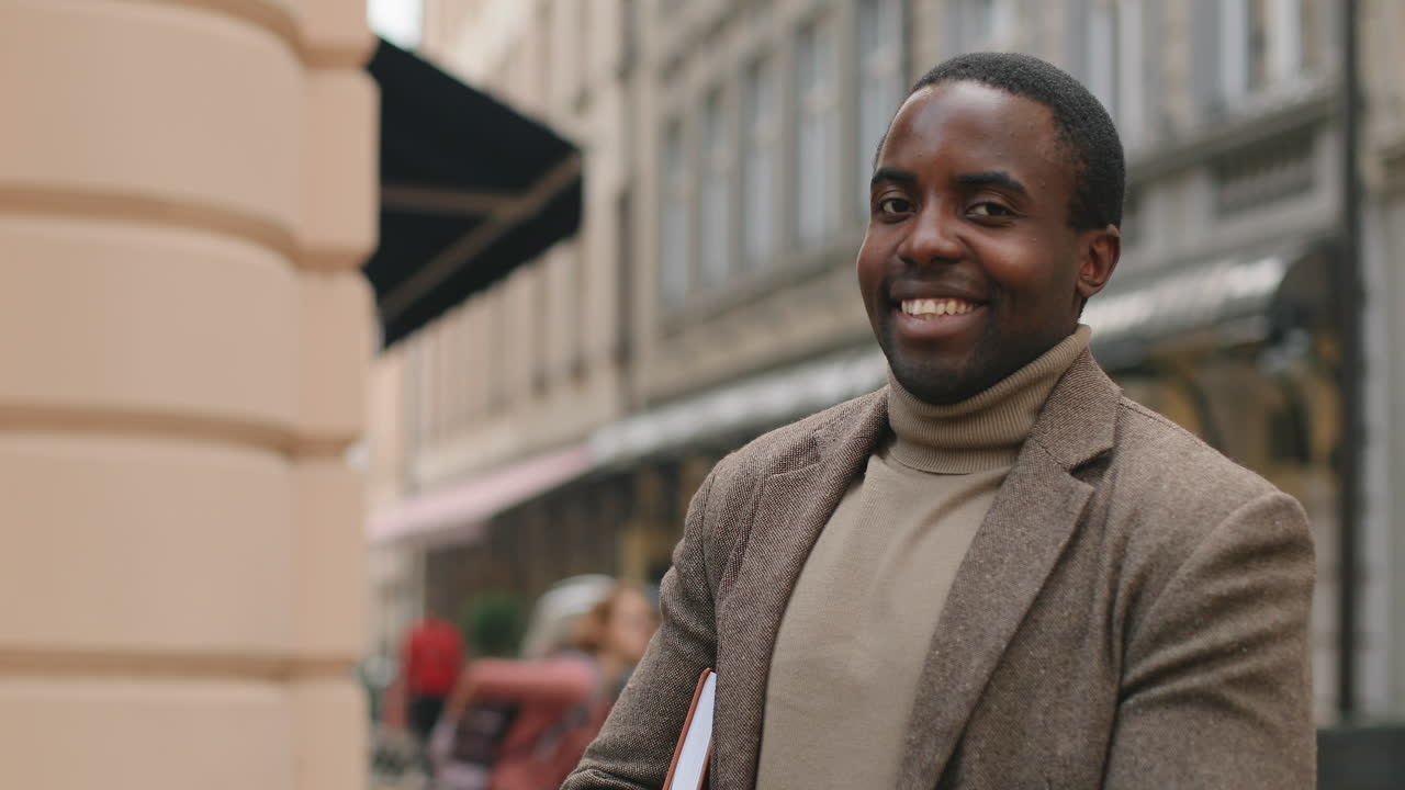 hombre de negocios afroamericano con traje elegante sosteniendo un cuaderno bajo el brazo, sonriendo a la cámara y caminando por la calle