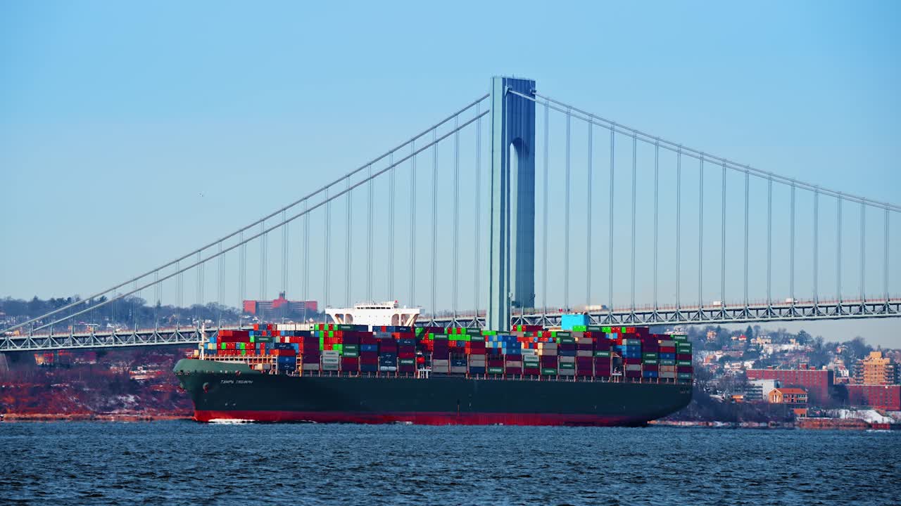 New York, USA, 5 October 2025: Container ship passing large bridge pylon at sunset. Green cargo vessel moving under a massive suspension bridge tower in golden hour light