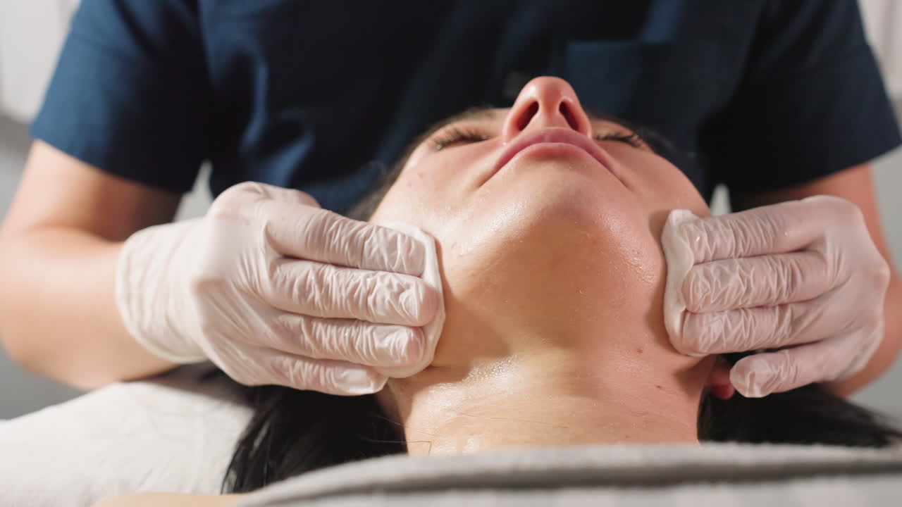 Close up view of skin care expert swabbing cotton pads across subject jawline to forehead during facial therapy session in bright spa clinic showcasing precision, calm and professional hygiene care