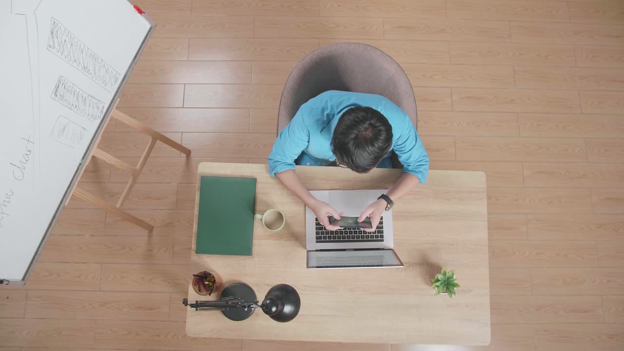 위쪽 뷰  아웃  한 남자가 집에서 노트북에서 타이핑하는 화이트보드 에 나무 책상에 앉아 스마트폰에서 게임을 하고 있습니다.