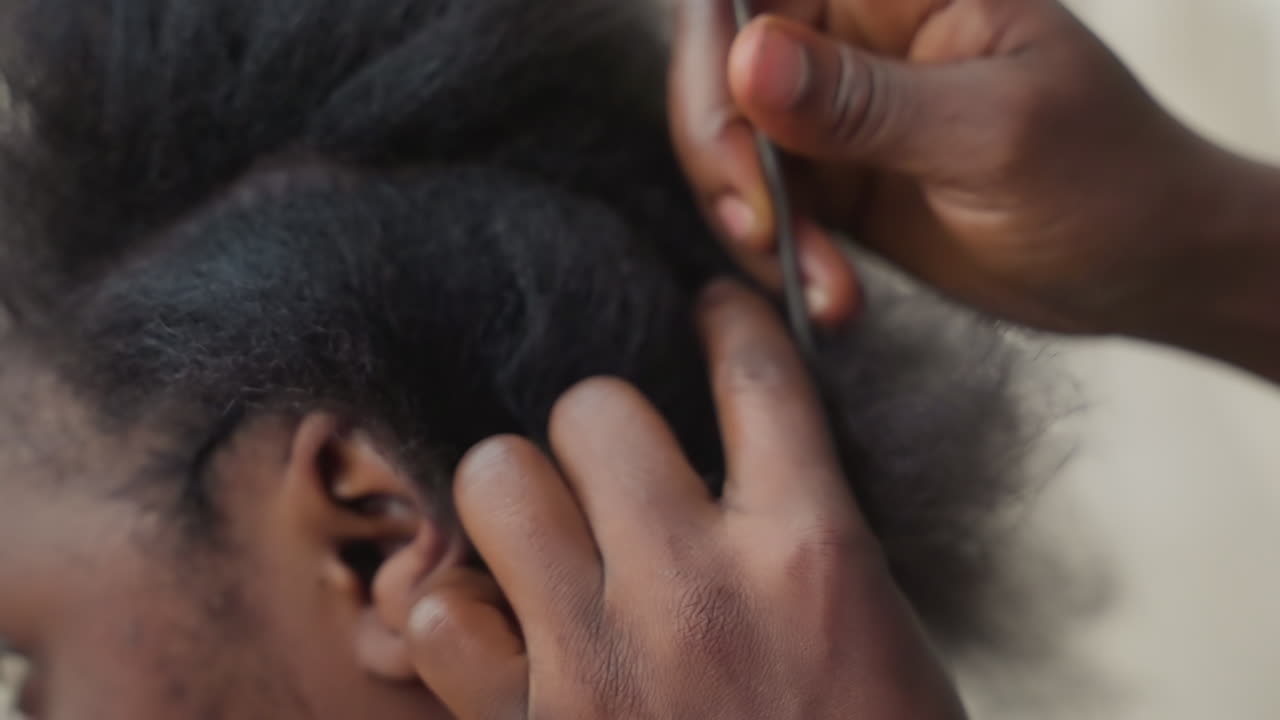 Specialista di trecce di colore che lavora da vicino su capelli naturali, dita che dividono e attorcigliano ciocche, pettine che solleva le radici, movimenti tattili precisi, cuoio capelluto texturizzato sotto una luce soffusa da studio, intensa concentrazione