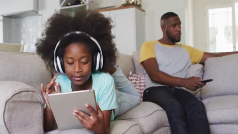hija afroamericana con auriculares usando una tableta mientras su padre mira la televisión