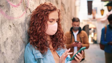 vista de cerca de una mujer pelirroja con máscara y usando un teléfono inteligente apoyado en una pared en la calle