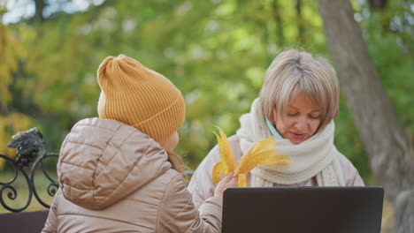 close up of woman sitting on bench working on laptop outdoors in autumn park while daughter offers yellow leaves to distract mother who gently shifts hand aside before child withdraws