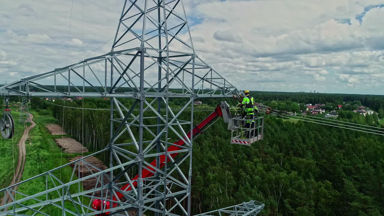 Two workers on a crane fixing a high-voltage tower in a wooded setting