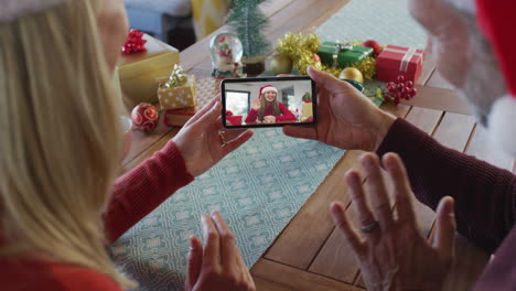 pareja caucásica con sombreros de santa usando un teléfono inteligente para una videollamada de navidad con una mujer en la pantalla