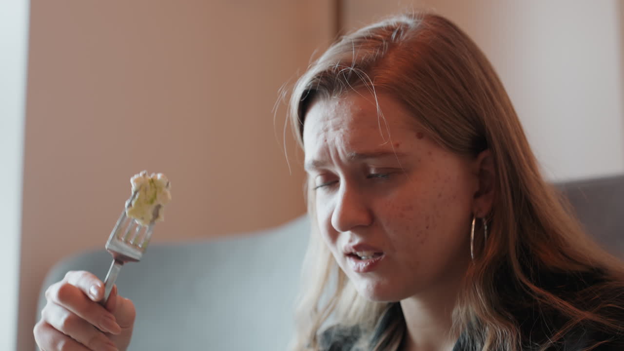 Hvid kvinde laver en grimasse ved en mundfuld, skuffet reaktion og eftertænksomt udtryk, indendørs sofa-portræt med dæmpet lys, gnider tindingen og viser træthed, madtester og sundhedsblogger evaluerer.