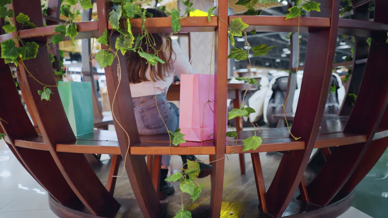 장식 된 나무 좌석에 앉아있는 젊은 성인, 에 전화, 쇼핑 가방을 들고, 사람들은 역동적인 조명과 함께 활기차고 바쁜 쇼핑몰에서 돌아다니는 것을 볼 수 있습니다.