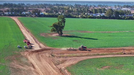 Vista-Aérea-De-Un-Tractor-Y-Una-Niveladora-Pasando-Junto-A-Otros-Vehículos-Agrícolas-A-Lo-Largo-De-Un-Camino-De-Tierra