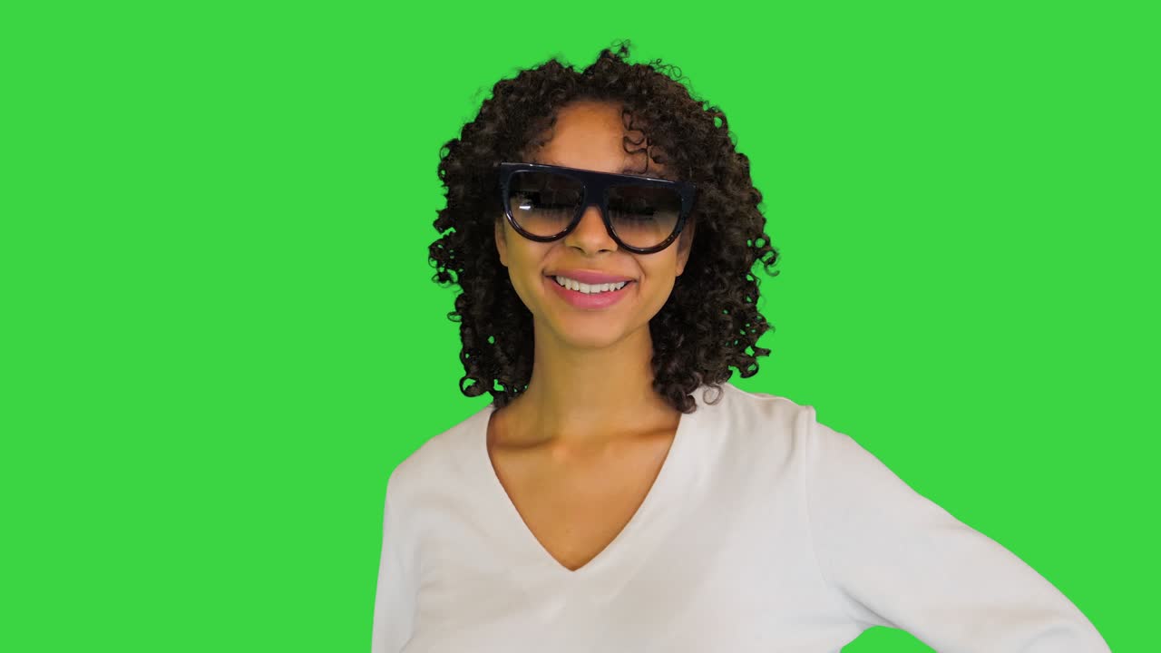 joven alegre poniendo gafas de sol y sonriendo a la cámara en una pantalla verde, clave de croma
