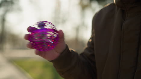 a close-up of an adult holding a purple toy with a rotating fan inside, the background shows a blurred view of trees and a walkway