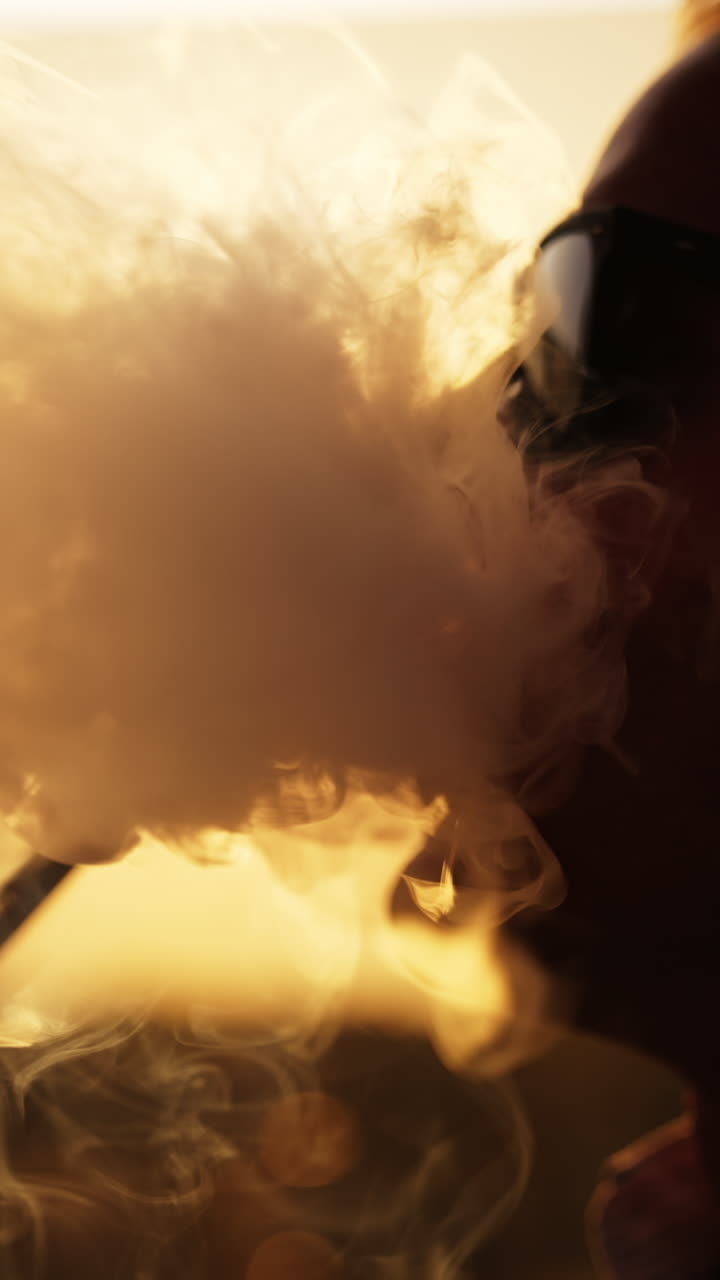 Silhouette of man in glasses smoking hookah sitting outdoors. Close up. Backdrop of setting sun. Vertical video