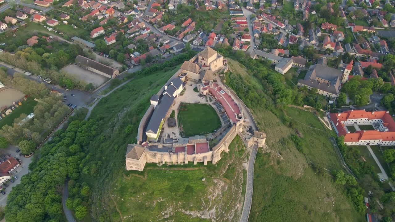 An aerial view of a historic medieval castle situated atop a green hill, surrounded by a quaint Hungarian town. The fortress showcases preserved stone walls, a courtyard, and traditional architecture
