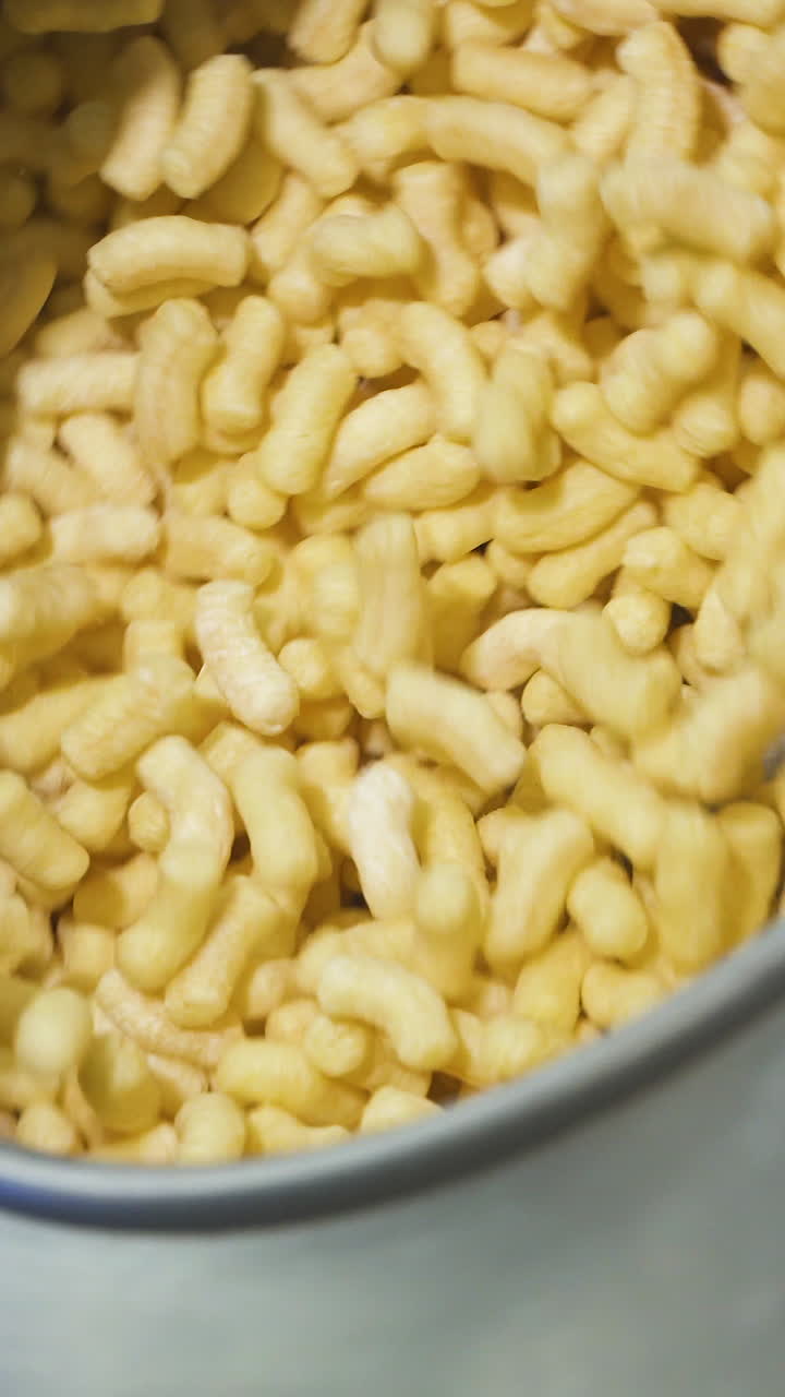 Rotating metal container with crispy corn sticks snacks. Tank for mixing products in a food factory. Vertical video