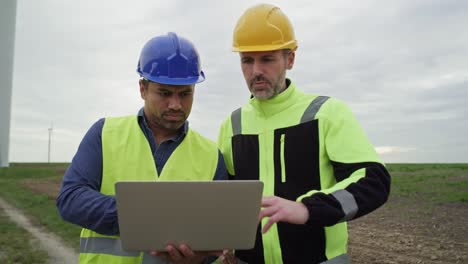 caucasian and latin male engineers standing on wind turbine field and discussing over computer.