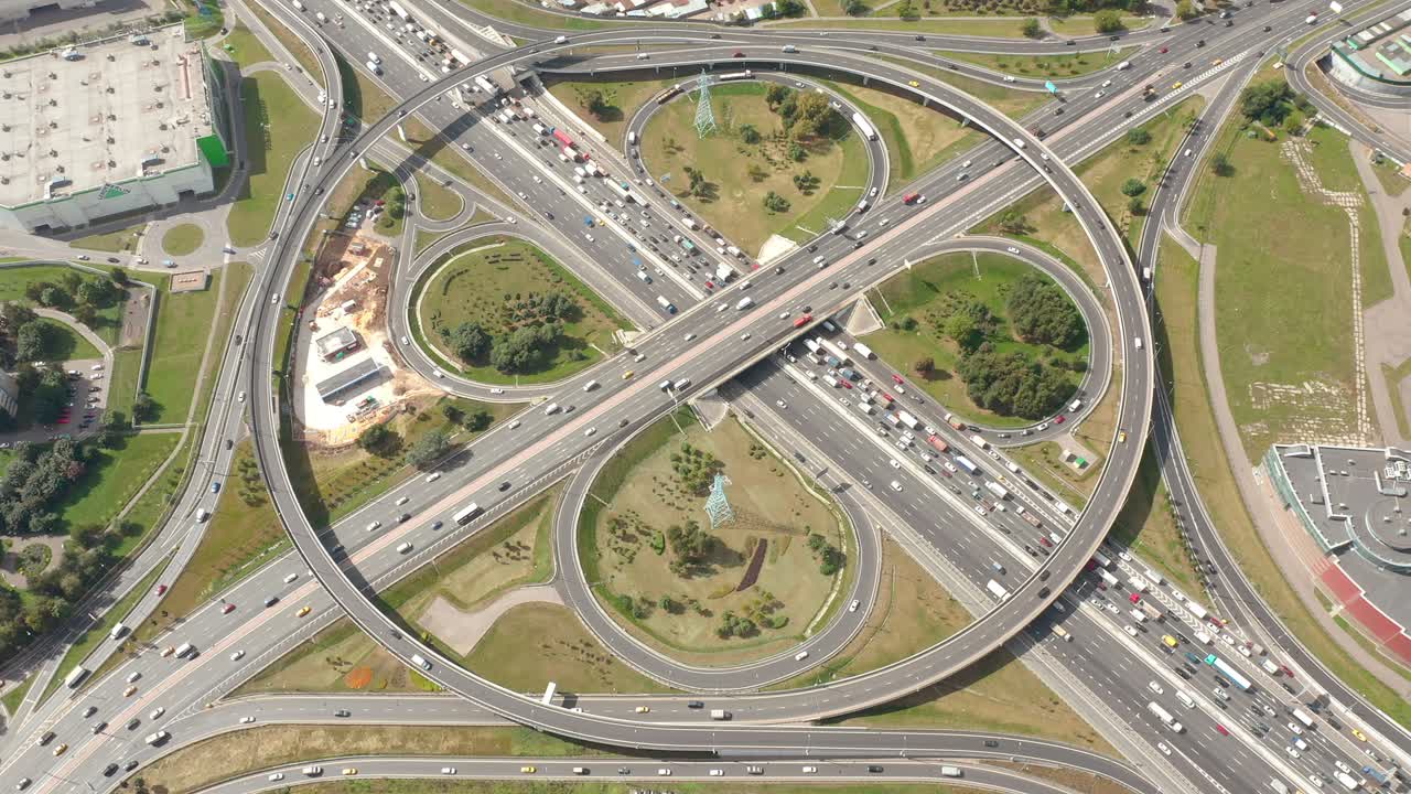vista estática en la gran intersección circular con vueltas, curvas y salidas de trébol. vista aérea superior desde un avión no tripulado. tráfico de la gran ciudad. luz soleada. plantaciones verdes, hierba y edificios alrededor. cámara estática y mirando hacia abajo a los coches en movimiento.