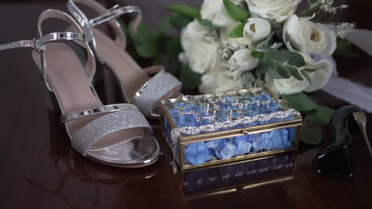 Close up of bridal accessories including silver heels, white bouquet, and ring box on a table